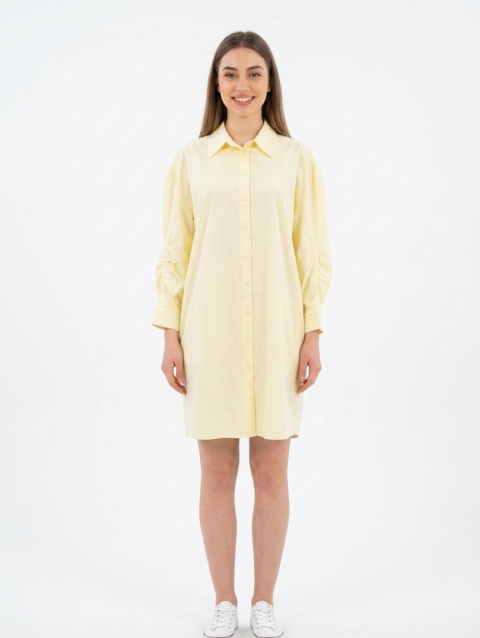 Mannequin souriante portant la robe chemise courte jaune pâle 'Aura' avec manches longues froncées et coupe ample, de la marque Divine Dress.
