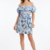 Mannequin portant la robe courte bleu clair à imprimé floral 'Bow Elegance', avec encolure Bardot à volants et grande ceinture nouée, de la marque Divine Dress